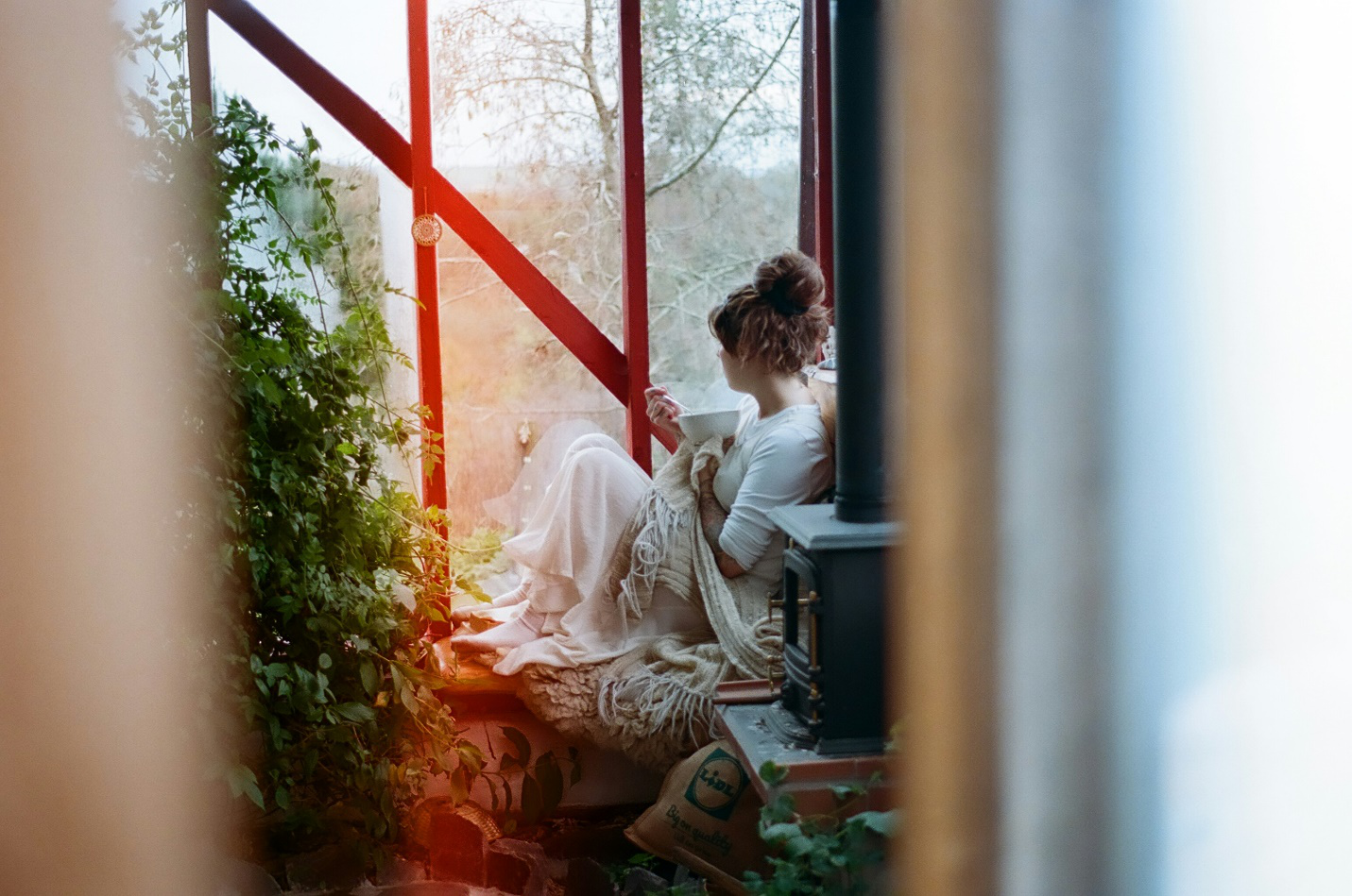 A woman in off-white sits with a bowl bundled up on a bench by a window with plants and shrubbery